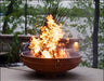 A round, rusted metal fire pit with a large, roaring fire burning inside. The fire pit has two handles on the side and is placed on a stone patio. There is a body of water and lush greenery in the background.