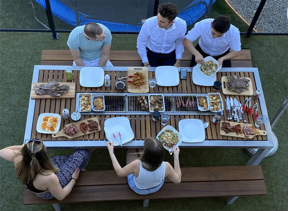 Group of people enjoying a meal outdoors on an ibbq table with various dishes.
