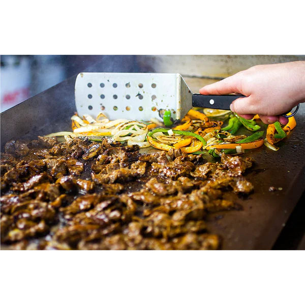 A hand using a spatula to stir a mixture of sliced peppers and onions cooking on a hot griddle. Sizzling meat is also visible on the griddle.