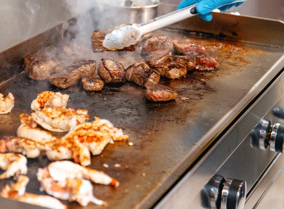 A close-up of a griddle with a variety of foods cooking on it. Steaks, chicken, and seafood are sizzling on the hot surface, with steam rising up.