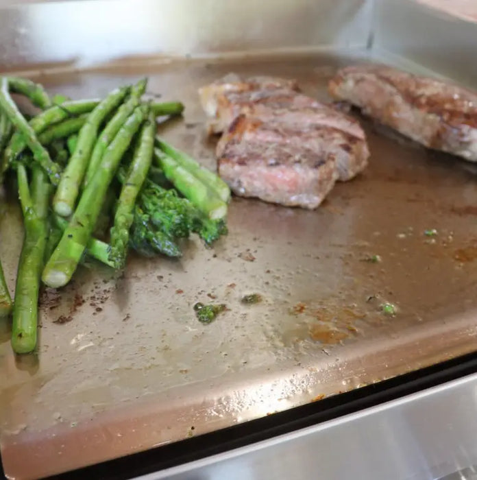 A close-up of a stainless steel griddle with steaks and asparagus cooking on it. The steaks have a nice sear and the asparagus is slightly charred.