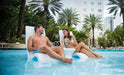 couple sitting on a simming pool chair inside the pool with a table next to them and enjoying drinks