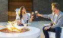 2 couples enjoying a a glass of red wine in outdoor living space around an outdoor propane fire pit. there is a stainless steel griddle in an outdoor kitchen behind them