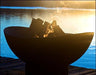  A round, black fire pit with flames burning inside. The fire pit is placed on a wooden dock near a body of water at sunset. The sky is orange and the water is reflecting the flames.