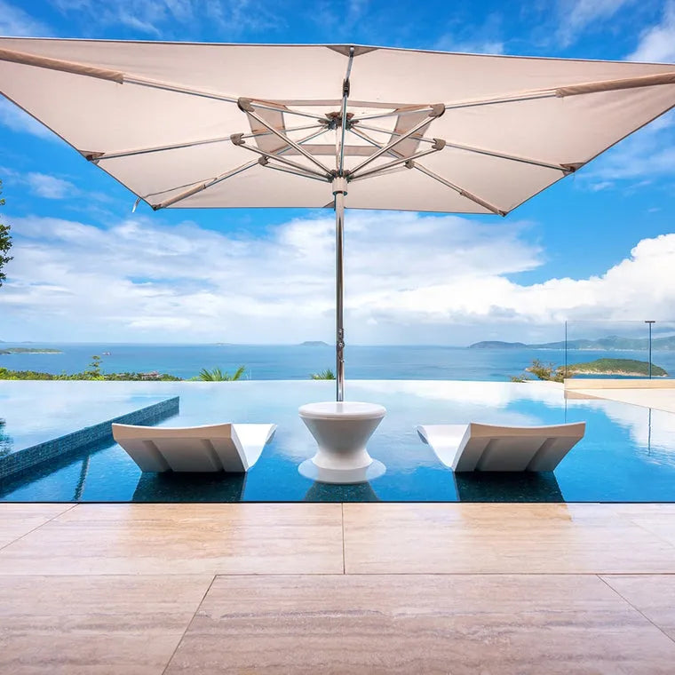 A spacious poolside area with a large, white cantilever umbrella providing shade. Two lounge chairs and a small table are positioned under the umbrella, inviting relaxation by the pool. The view stretches out to the ocean and distant hills.