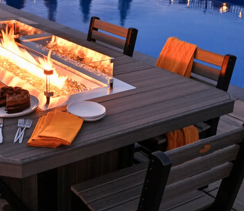Outdoor dining setup with fire pit, table, chairs, and cake by a pool at night.