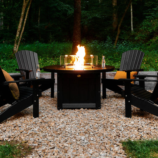 A 46-inch round fire pit table with a roaring fire, surrounded by four black Adirondack chairs on a gravel patio in a wooded backyard.