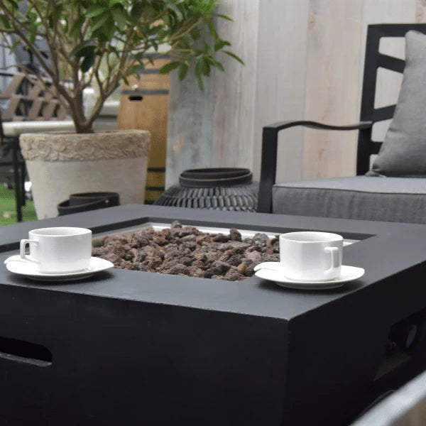 2 white cups of coffee cups on square fire pit table. there is a fire pit in the middle that has lava rocks laid but lava rocks are not lit.