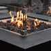 square fire pit with matching glass square windscreen. Fire pit is lit on lava rocks and yellow and orange flames is lit. Close up view of glass windscreen and square fire pit table