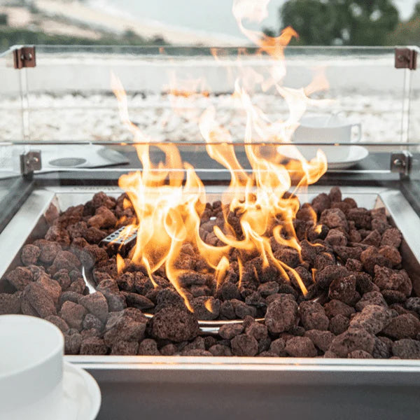 close of view of fire pit flame that is burning on dark brown lava rocks and has glass windscreen around it