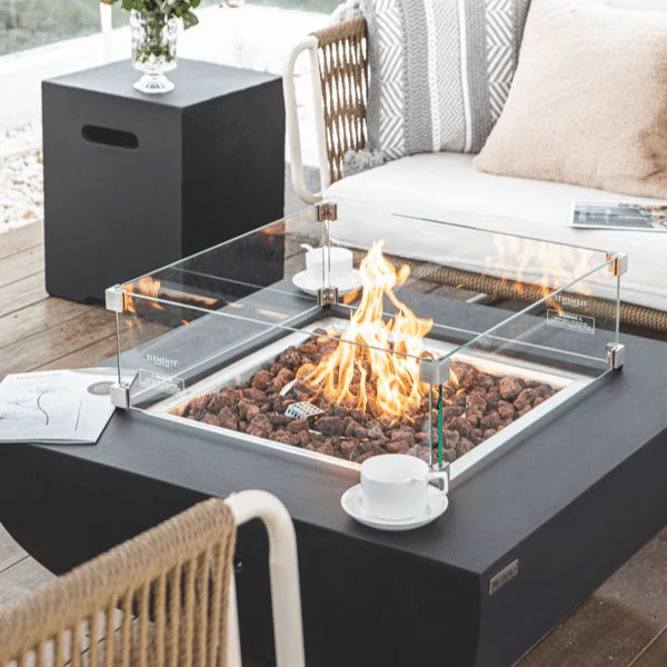 square fire pit table that is lit on lava rocks. It has a matching square glass windscreen and a coffee cup and magazine sitting on the side of the fire table. there is a matching tank cover that is being used as a table top to is holding a flower vase