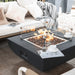 square fire pit table is lit on lava rocks with glass windscreen  and 2 cups of coffee on the side of the table  with outdoor sofa next to it