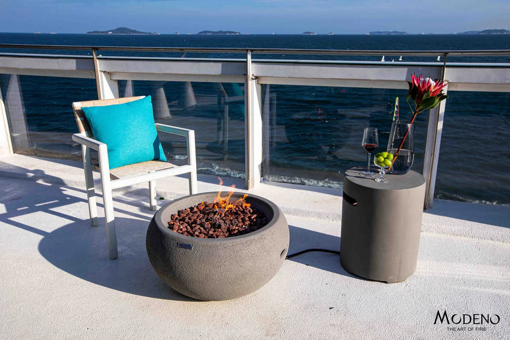 round fire pit bowl lit on lava rocks and connected to matching smooth top tank cover on a balcony overlooking the sea