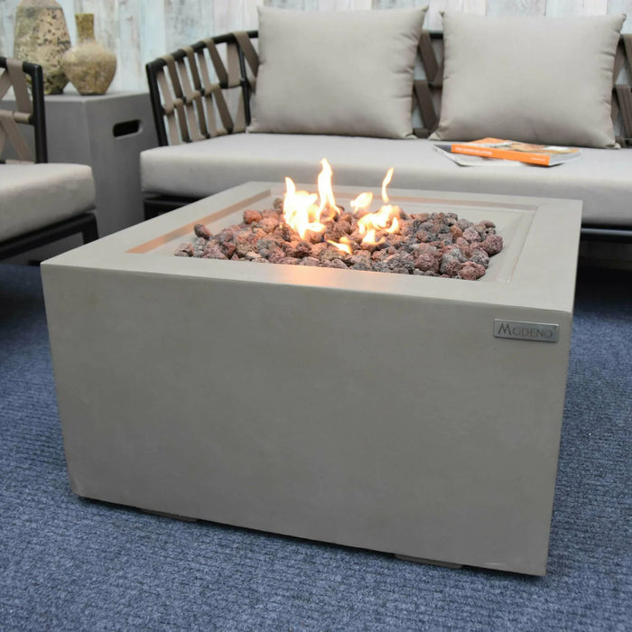 light grey square fire pit table with lava rocks by modeno. lava rocks are lit and flames are yellow and organce. fire pit table is in the middle of furniture