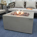 light grey square fire pit table with lava rocks by modeno. lava rocks are lit and flames are yellow and organce. fire pit table is in the middle of furniture