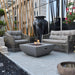 light grey square fire pit table in front of outdoor seating in a backyard. fire pit is lit on lava rocks