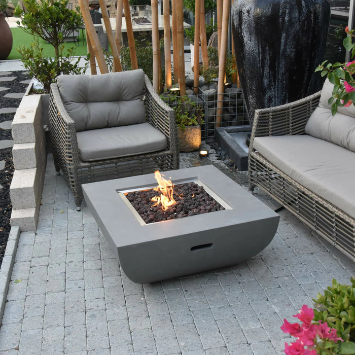 light grey square fire pit table lit on lava rocks next to outdoor furniture in a backyard