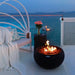 black round fire pit is lit on lava rocks on a deck overlooking the sea. It has a matching tank cover beside it that is acting like a table told to hold a vase of flowers