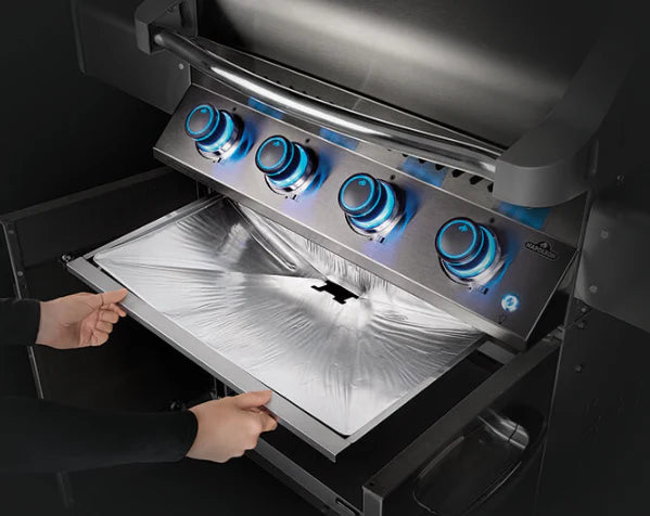 A person removing a silver aluminum foil drip pan liner from a stainless steel gas grill by napoleon grills on their pro prestige 500 model