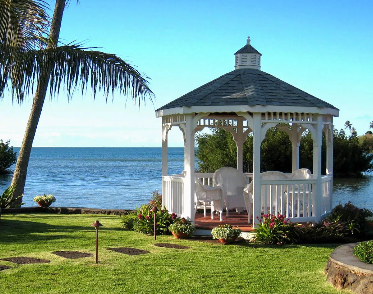 White gazebo with fire pit and chairs on a grassy area by a body of water with palm trees.