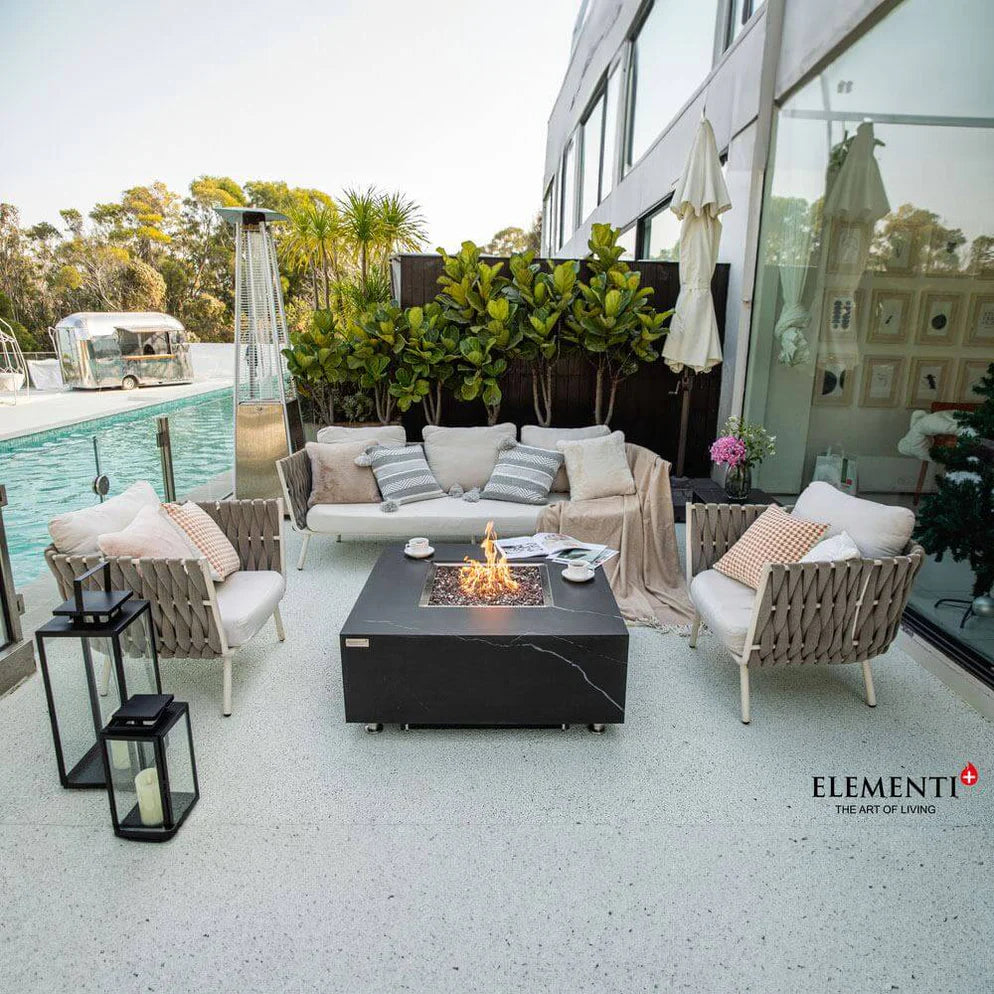 Modern outdoor patio featuring a square black fire pit table with a glass surround, surrounded by a white sofa and chairs with light cushions. The patio overlooks a swimming pool and lush greenery with a glimpse of a vintage trailer.