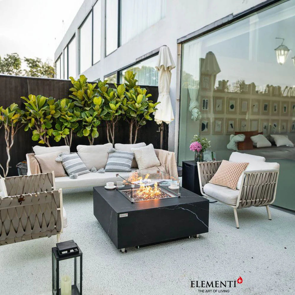 Contemporary outdoor patio featuring a square black fire pit table with a glass surround, surrounded by a white sofa and chairs with light cushions. The patio has a modern design with large windows and overlooks a leafy green garden.