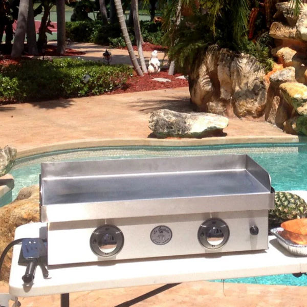 A large outdoor griddle that is stainless steel and sits on a white table outdoors. The griddle has three round knobs and a power cord. In the background, there is a swimming pool and lush landscaping.