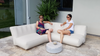 Two women sitting on a modern outdoor sofa with a small table between them, surrounded by plants.
