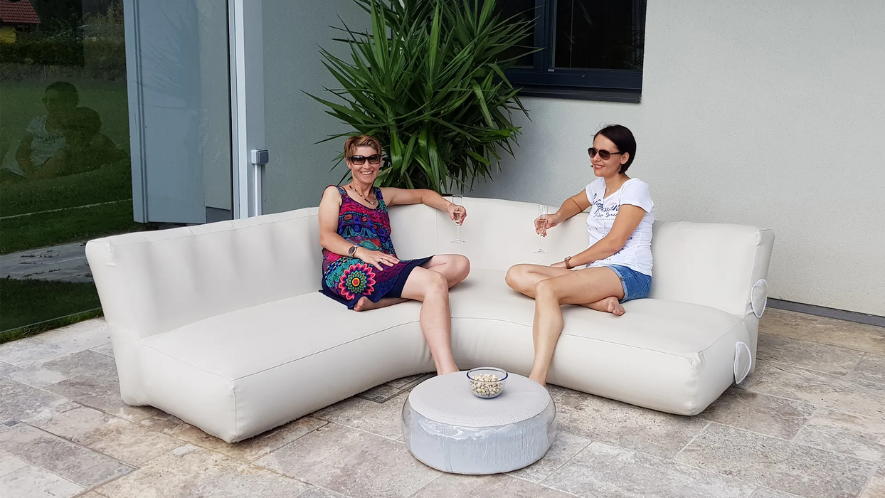 Two women sitting on a modern outdoor sofa with a small table between them, surrounded by plants.