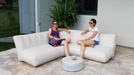 Two women sitting on a modern outdoor sofa with a small table between them, surrounded by plants.