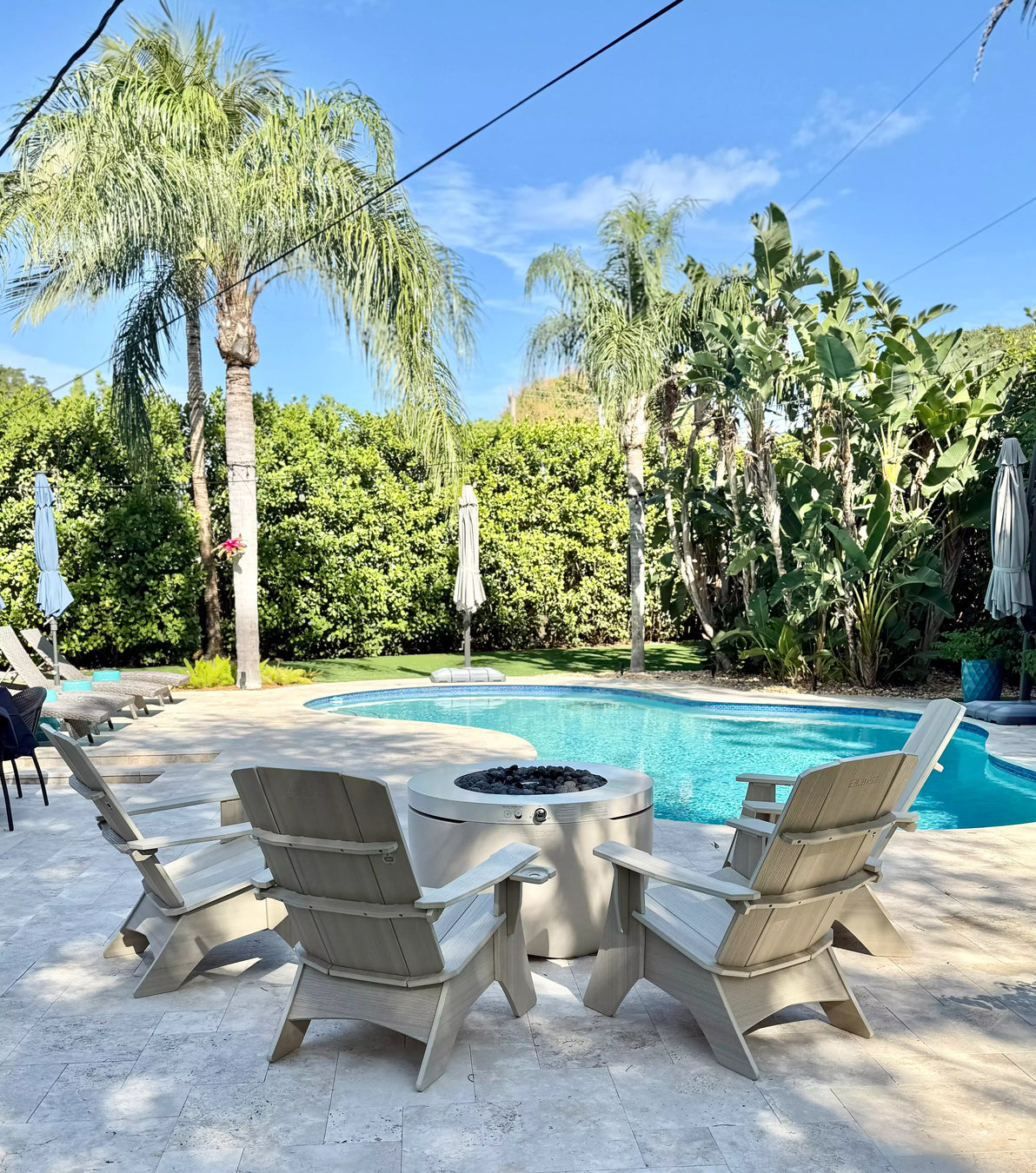 Stone patio with a central fire pit and comfortable seating, with a blue swimming pool, palm trees, and various plants in the background.