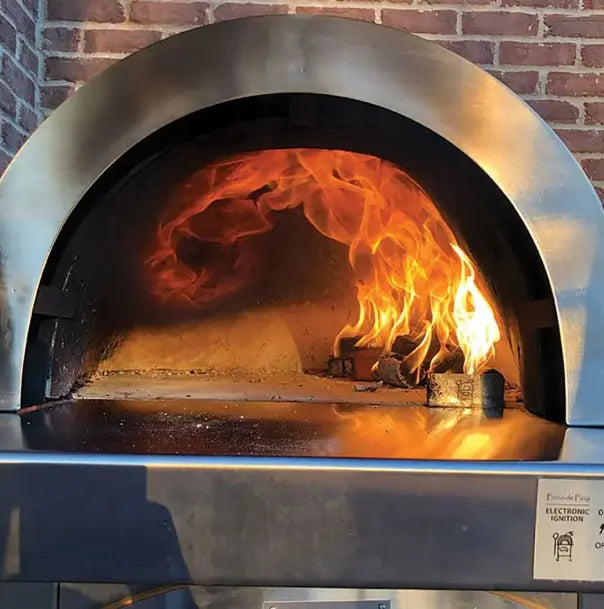 A vintage cast iron pizza oven with a red finish. The oven has a glass window in the door and a metal chimney on top. It sits on a metal stand with four legs.