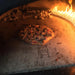 A round pizza with a browned crust is cooking in a brick oven. Flames are visible through the opening of the oven. The oven is made of light-colored bricks and has a metal archway over the opening.