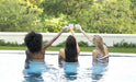 3 women saying cheers with their  glasses high in the air while they sit on in pool bar stool on swimming pool ledge