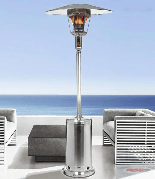 Stainless steel patio heater with a reflective heat shield and a propane tank base, positioned on a patio overlooking the ocean.