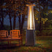 A silver pyramid-shaped propane patio heater with a woven base, casting a warm glow over a wooden bench and glasses on a table in a backyard at night.