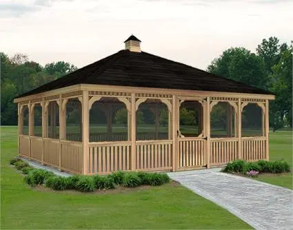 A large rectangular wooden gazebo with a peaked roof and screened walls. The gazebo features decorative latticework and a double door with a small window. It is surrounded by a paved walkway and a lawn with mature trees in the background.