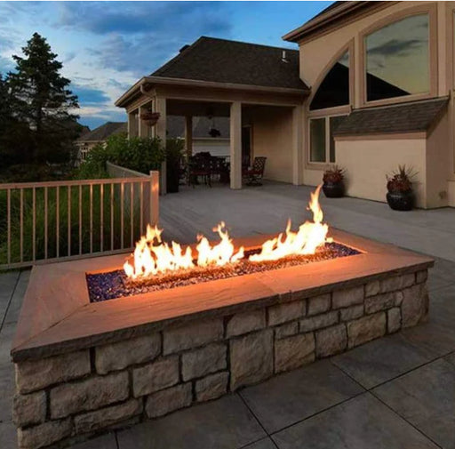 A rectangular fire pit with orange flames burning in it sits on a stone patio in front of a house with a brown exterior. There are a few trees in the background.