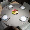 A high-angle, top-down shot captures a round dining table set for four. The table has a smooth, light grey surface. Four chairs, only partially visible around the edges of the frame, surround the table