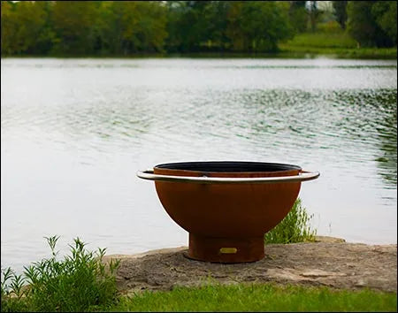 A round, rust-colored wood burning fire pit with a stainless steel handle. It is sitting on a rock near a lake with trees in the background.