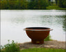 A round, rust-colored wood burning fire pit with a stainless steel handle. It is sitting on a rock near a lake with trees in the background.