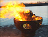 A round, rusted metal fire pit with flames burning inside. The fire pit has a seashell design cut into the metal. It is placed on a rock near a body of water at sunset.