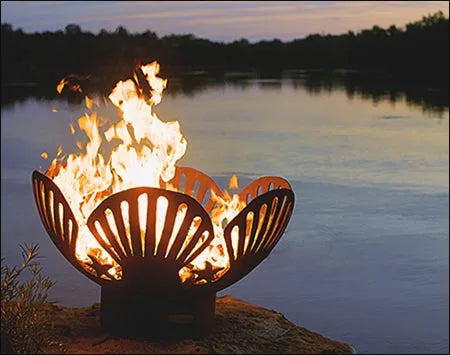 A wood-burning fire pit shaped like a seashell, with flames dancing within. The fire pit is placed on a rocky shoreline near a lake.