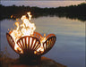 A wood-burning fire pit shaped like a seashell, with flames dancing within. The fire pit is placed on a rocky shoreline near a lake.