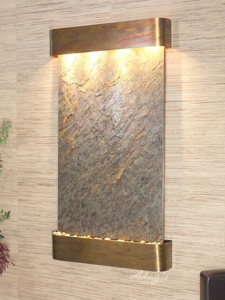 
A vertical, rectangular wall mounted water feature featuring a textured natural stone panel. The stone is a mix of grays and browns with visible variations and ridges. At the top, a horizontal, U-shaped metal hood in a warm, possibly copper or brass, finish contains multiple lights that illuminate the stone surface below.