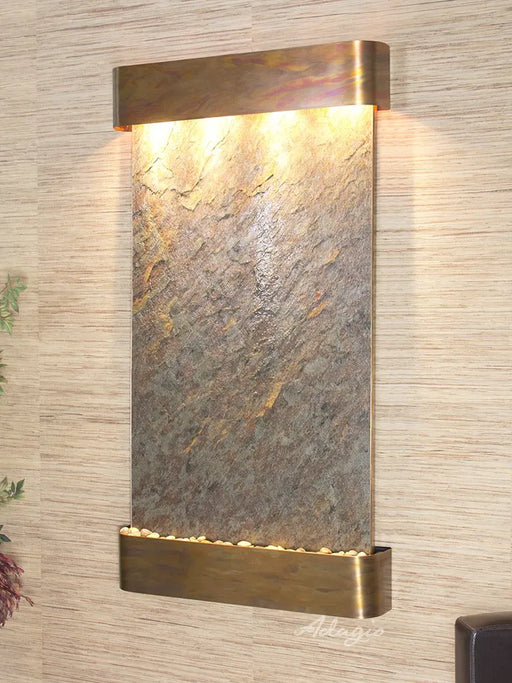 
A vertical, rectangular wall mounted water feature featuring a textured natural stone panel. The stone is a mix of grays and browns with visible variations and ridges. At the top, a horizontal, U-shaped metal hood in a warm, possibly copper or brass, finish contains multiple lights that illuminate the stone surface below.