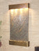 
A vertical, rectangular wall mounted water feature featuring a textured natural stone panel. The stone is a mix of grays and browns with visible variations and ridges. At the top, a horizontal, U-shaped metal hood in a warm, possibly copper or brass, finish contains multiple lights that illuminate the stone surface below.