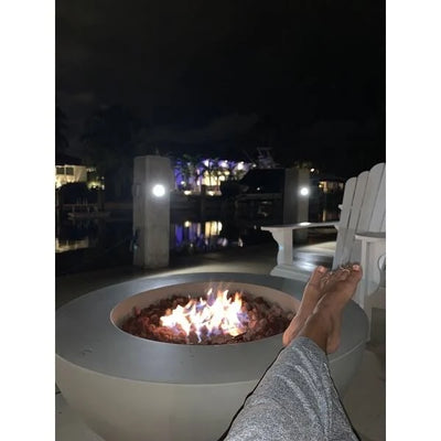 lady perching feet on elementi luna fire pit bowl that has got round smooth edges and it lit on a bed of red lava rocks