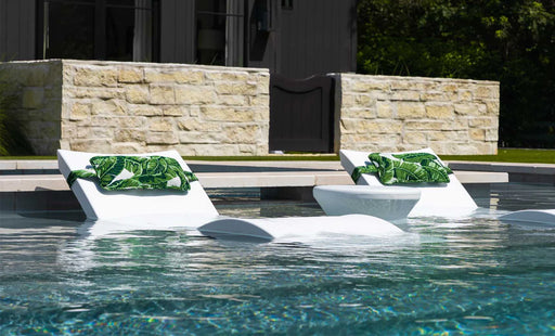 Two white in-pool chaise lounge chairs with green tropical leaf patterned pillows, positioned on a tanning ledge in a modern pool with stone retaining walls.