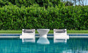 Two white modern pool loungers with gray cushions sit in a shallow pool area, facing a small white table. A neatly trimmed green hedge is in the background.